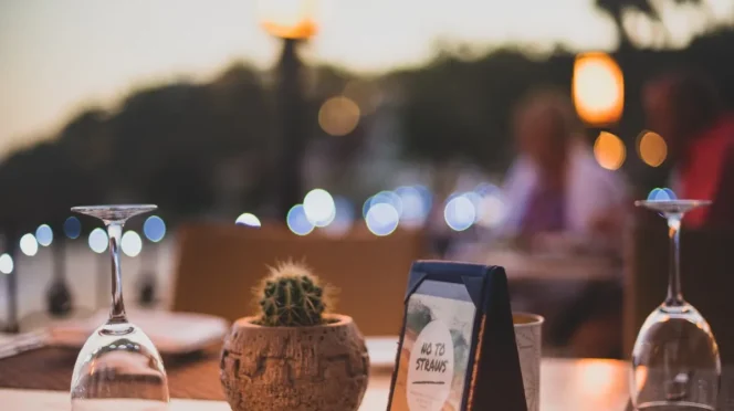 Table setting at a restaurant with a small cactus in a clay pot, a framed 'No To Straws' sign, and two inverted wine glasses; warm bokeh lights in the background.