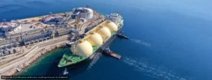 Aerial view of a large LNG tanker docked at a gas terminal, with four beige spherical storage tanks on the deck and industrial facilities onshore amid blue ocean.