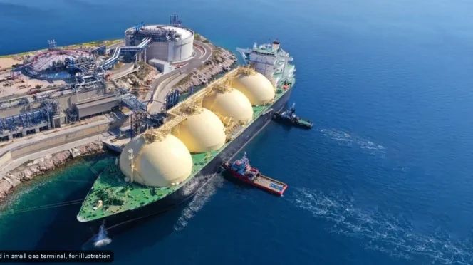 Aerial view of a large LNG tanker docked at a gas terminal, with four beige spherical storage tanks on the deck and industrial facilities onshore amid blue ocean.