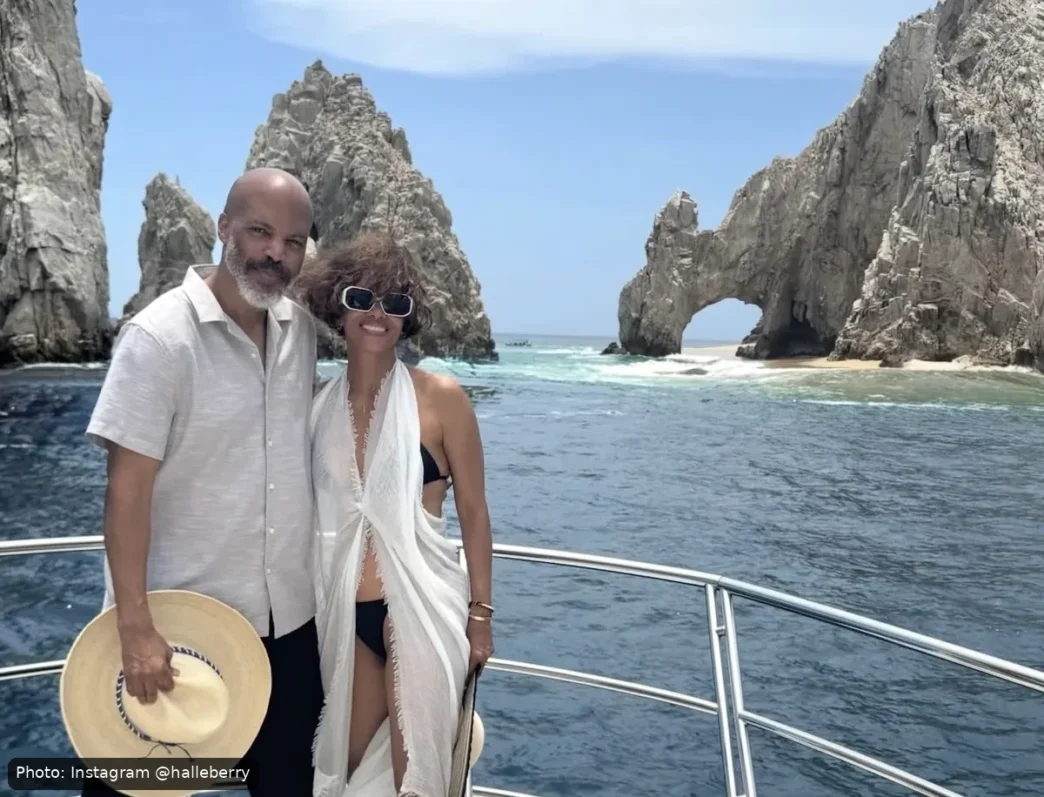 Couple posing on a boat with a rocky sea arch and blue ocean behind them under a clear sky.