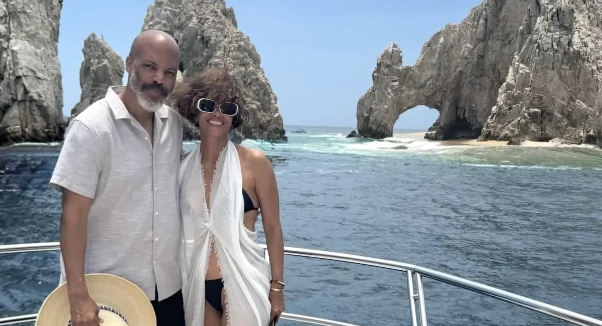 Couple posing on a boat with a rocky sea arch and blue ocean behind them under a clear sky.