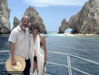 Couple posing on a boat with a rocky sea arch and blue ocean behind them under a clear sky.