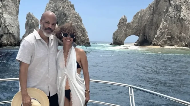 Couple posing on a boat with a rocky sea arch and blue ocean behind them under a clear sky.