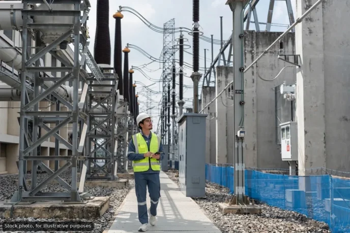 Utility worker in a hard hat and high-visibility vest walks between high‑voltage substation equipment and transformers.