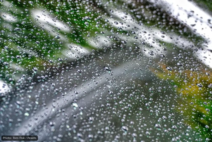 Close-up of raindrops on a car windshield with a blurred street and greenery in the background.