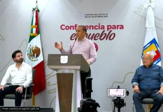 Speaker at podium delivering a press conference, with the Baja California Sur flag and a banner reading 'Conferencia para el pueblo' in the background.
