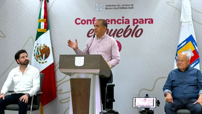 Speaker at podium delivering a press conference, with the Baja California Sur flag and a banner reading 'Conferencia para el pueblo' in the background.