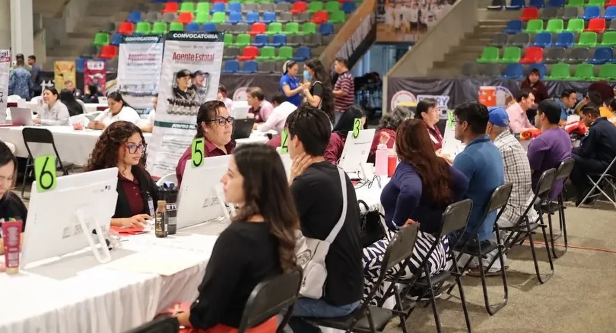 People sit at long white tables with computers and green numbered signs (5, 6) at a job fair inside a large arena with colorful bleachers in the background; staff assist applicants at each station.