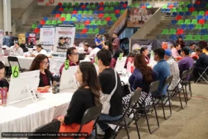 People sit at long white tables with computers and green numbered signs (5, 6) at a job fair inside a large arena with colorful bleachers in the background; staff assist applicants at each station.