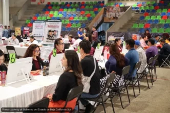 People sit at long white tables with computers and green numbered signs (5, 6) at a job fair inside a large arena with colorful bleachers in the background; staff assist applicants at each station.
