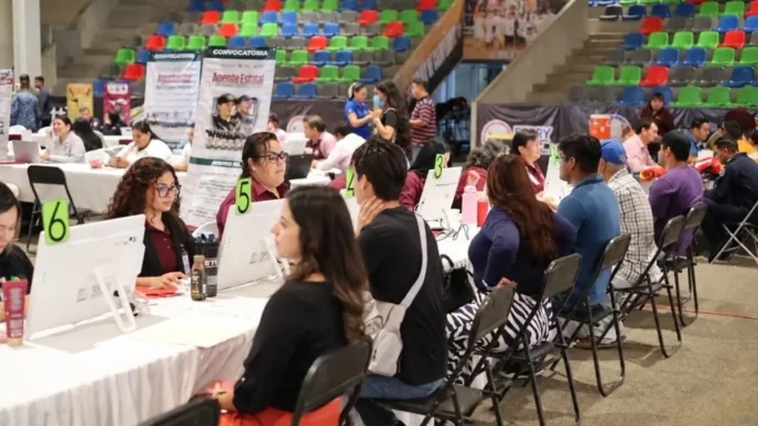 People sit at long white tables with computers and green numbered signs (5, 6) at a job fair inside a large arena with colorful bleachers in the background; staff assist applicants at each station.