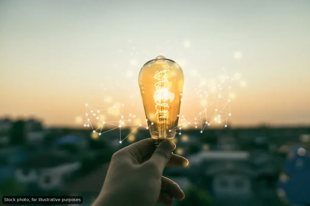 A hand holding a lit lightbulb with digital network lines in the air, symbolizing ideas and innovation at sunset.