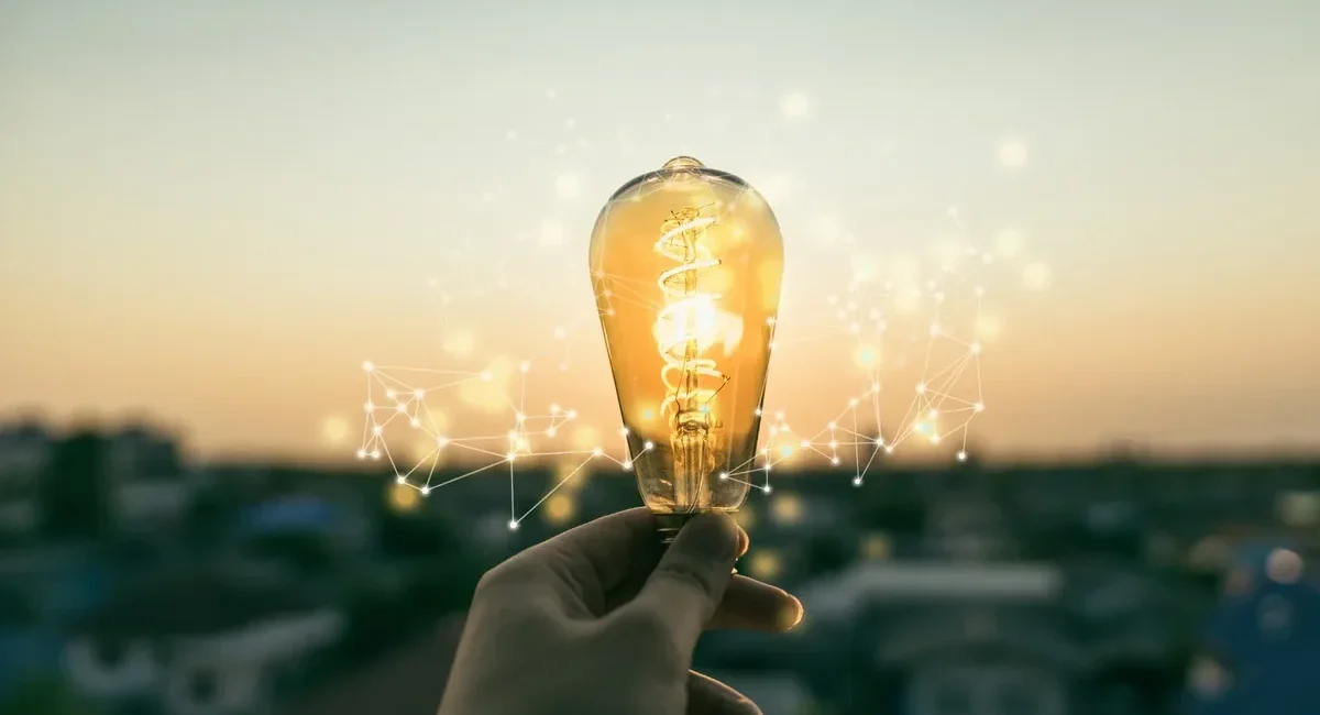 A hand holding a lit lightbulb with digital network lines in the air, symbolizing ideas and innovation at sunset.