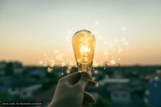 A hand holding a lit lightbulb with digital network lines in the air, symbolizing ideas and innovation at sunset.