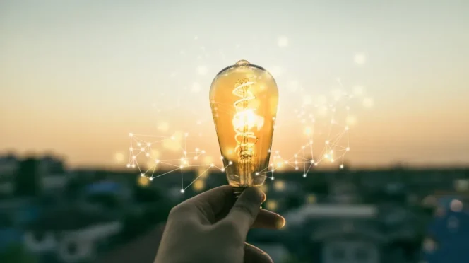 A hand holding a lit lightbulb with digital network lines in the air, symbolizing ideas and innovation at sunset.