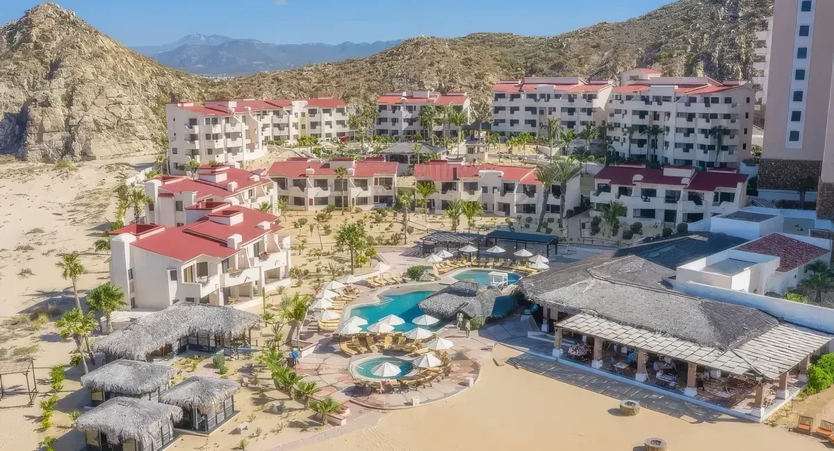 Beachfront resort with white buildings and red roofs surrounding a central pool area with circular pools and thatched palapas.