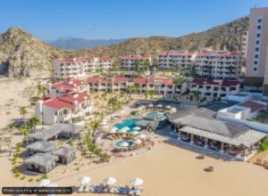 Beachfront resort with white buildings and red roofs surrounding a central pool area with circular pools and thatched palapas.