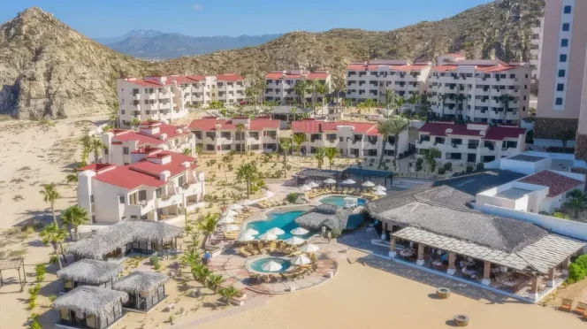 Beachfront resort with white buildings and red roofs surrounding a central pool area with circular pools and thatched palapas.
