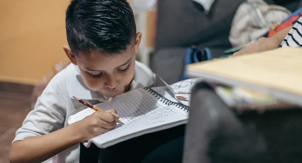 Young boy sitting on the floor, writing in a spiral notebook with a pencil, concentrating on homework at home