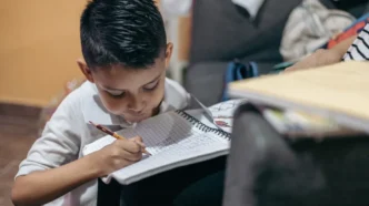Young boy sitting on the floor, writing in a spiral notebook with a pencil, concentrating on homework at home