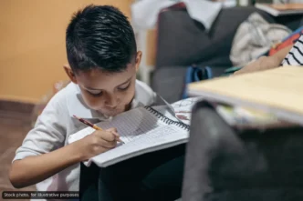 Young boy sitting on the floor, writing in a spiral notebook with a pencil, concentrating on homework at home
