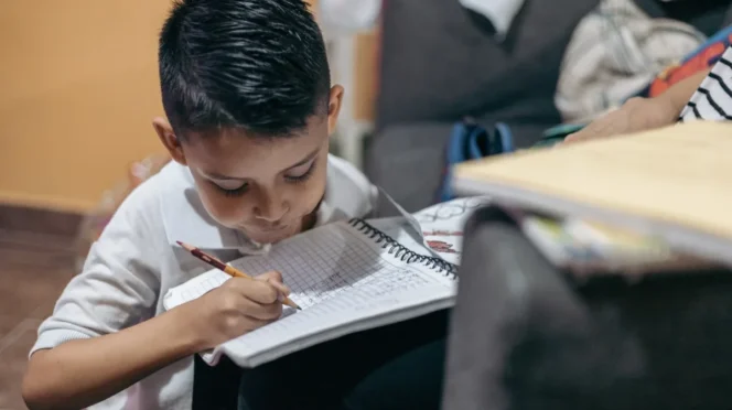 Young boy sitting on the floor, writing in a spiral notebook with a pencil, concentrating on homework at home