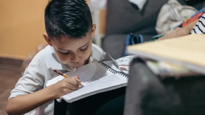 Young boy sitting on the floor, writing in a spiral notebook with a pencil, concentrating on homework at home