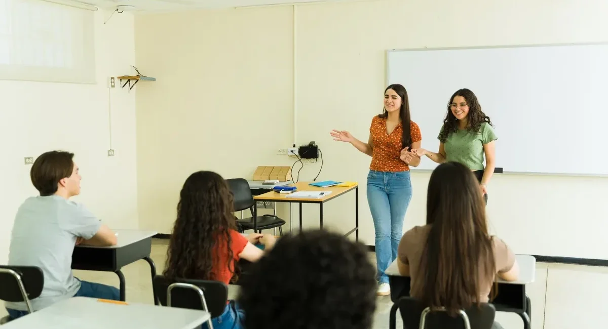 Two young women present at the front of a classroom while peers sit at desks and listen near a whiteboard.