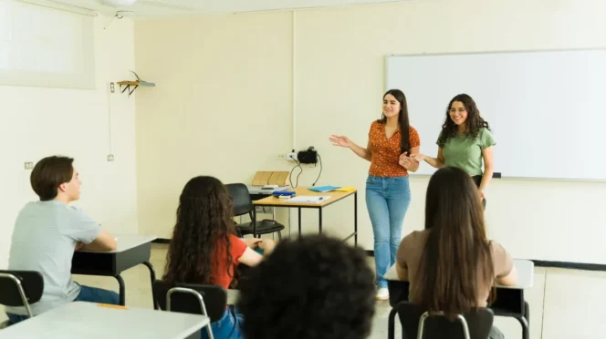 Two young women present at the front of a classroom while peers sit at desks and listen near a whiteboard.