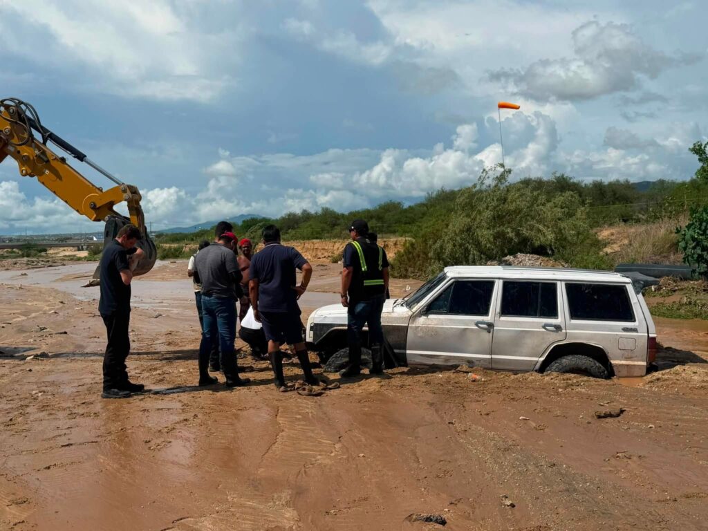 Group of workers at a muddy construction site examining a yellow excavator arm beside a parked white SUV. An orange windsock rises in the background.