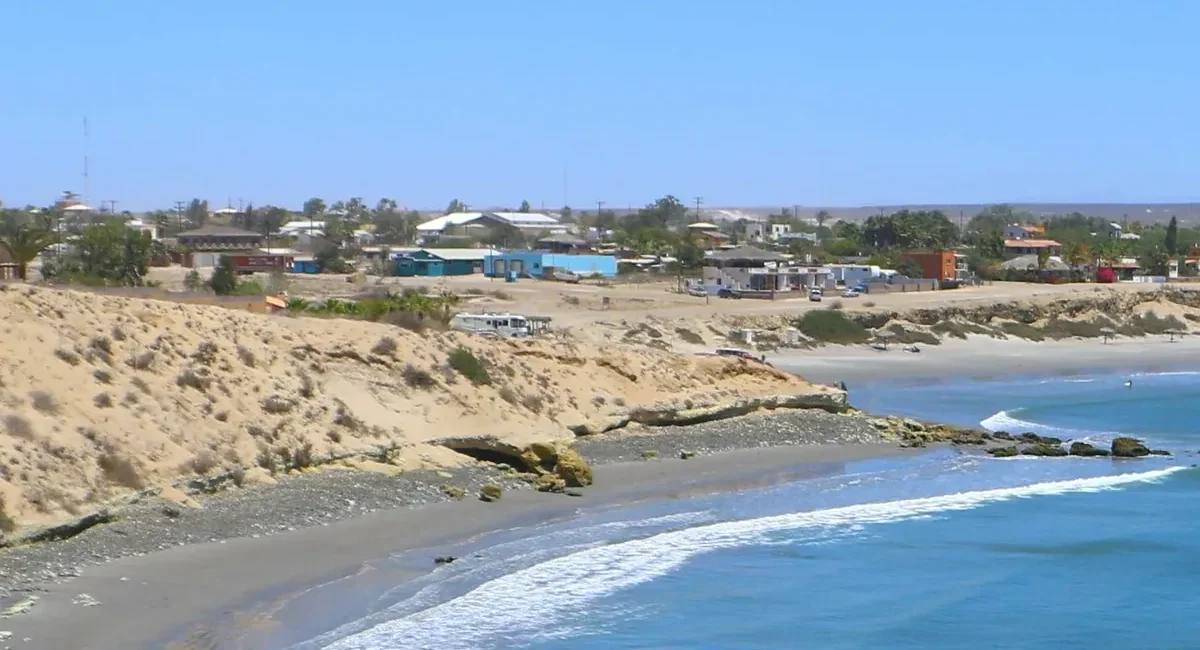 Coastal town with sandy cliffs, colorful houses, and a calm blue sea along a pebble beach.