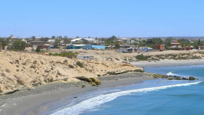 Coastal town with sandy cliffs, colorful houses, and a calm blue sea along a pebble beach.