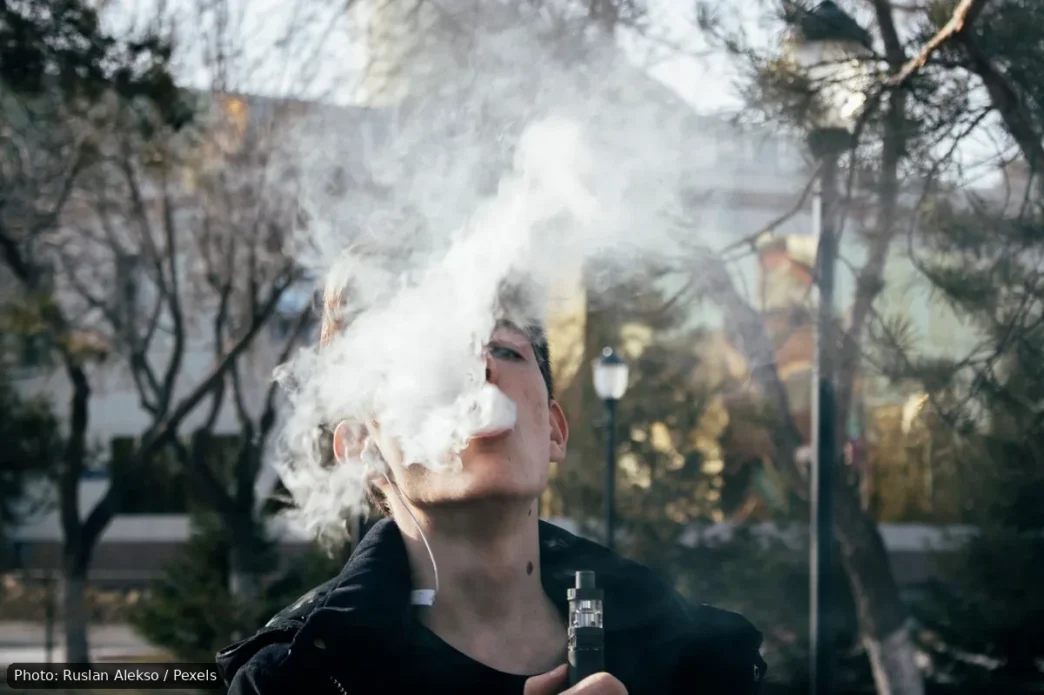 Young man vaping outdoors, blowing a large cloud of vapor toward the camera.