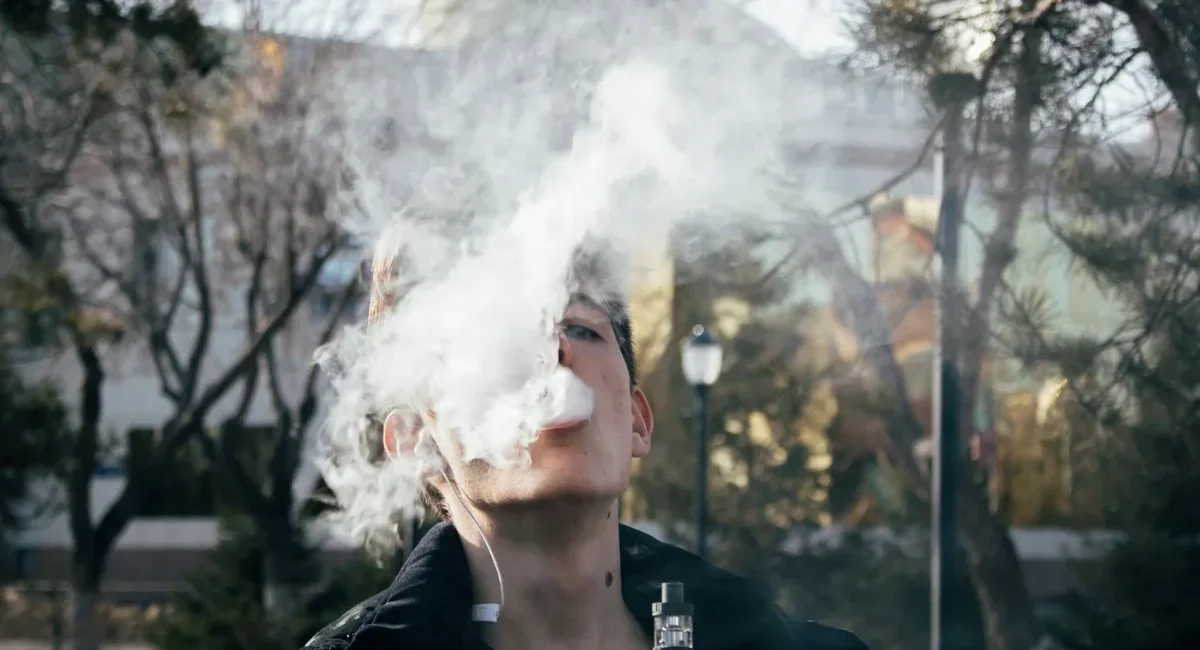 Young man vaping outdoors, blowing a large cloud of vapor toward the camera.