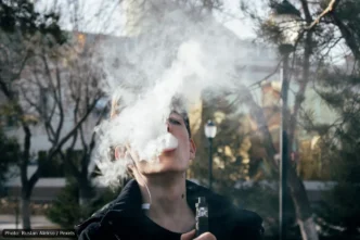 Young man vaping outdoors, blowing a large cloud of vapor toward the camera.