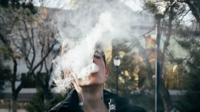 Young man vaping outdoors, blowing a large cloud of vapor toward the camera.