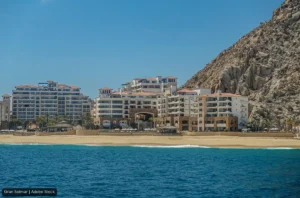 Resort buildings line a sandy beach with a rocky cliff to the right and calm blue sea in the foreground.