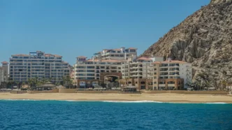 Resort buildings line a sandy beach with a rocky cliff to the right and calm blue sea in the foreground.