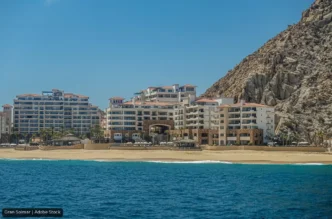 Resort buildings line a sandy beach with a rocky cliff to the right and calm blue sea in the foreground.
