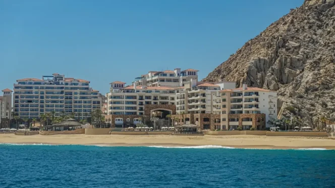 Resort buildings line a sandy beach with a rocky cliff to the right and calm blue sea in the foreground.