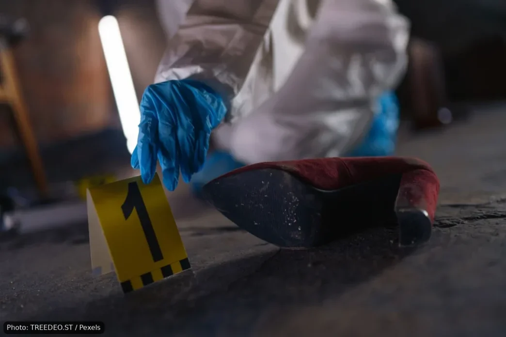 Forensic investigator in blue gloves places a yellow evidence marker labeled 1 beside a red high-heeled shoe on a dusty floor.
