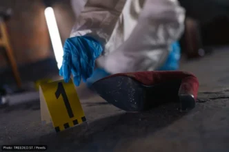 Forensic investigator in blue gloves places a yellow evidence marker labeled 1 beside a red high-heeled shoe on a dusty floor.