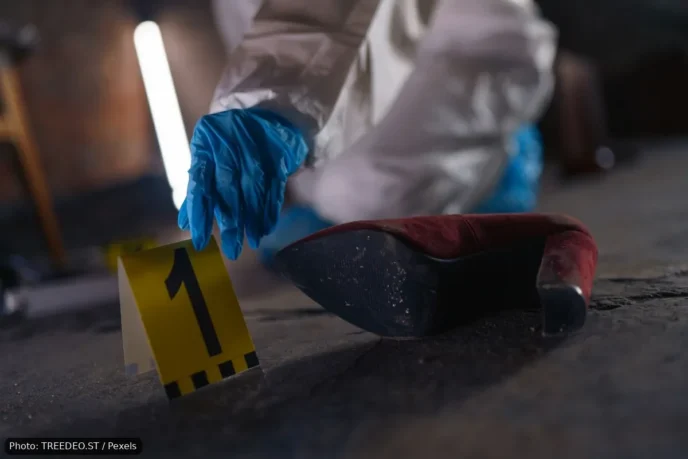 Forensic investigator in blue gloves places a yellow evidence marker labeled 1 beside a red high-heeled shoe on a dusty floor.