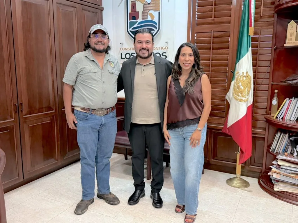 Three people stand together in a formal office, smiling for the photo, with a Mexican flag nearby and a wooden backdrop.