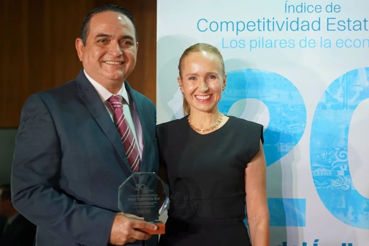 BCS Ranks 5th in National Competitiveness Index - Gringo Gazette Man in a dark suit holding a glass award, standing beside a smiling woman in a black dress at an awards event backdrop.