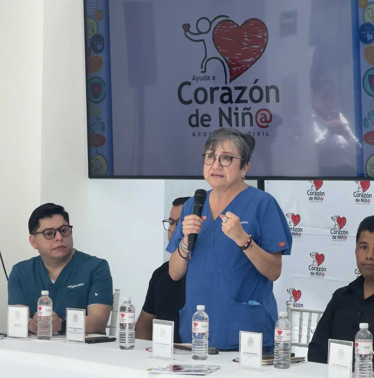 Woman in blue scrubs stands and speaks into a microphone at a panel for Corazón de Niñ@, with a heart logo on the backdrop behind her.