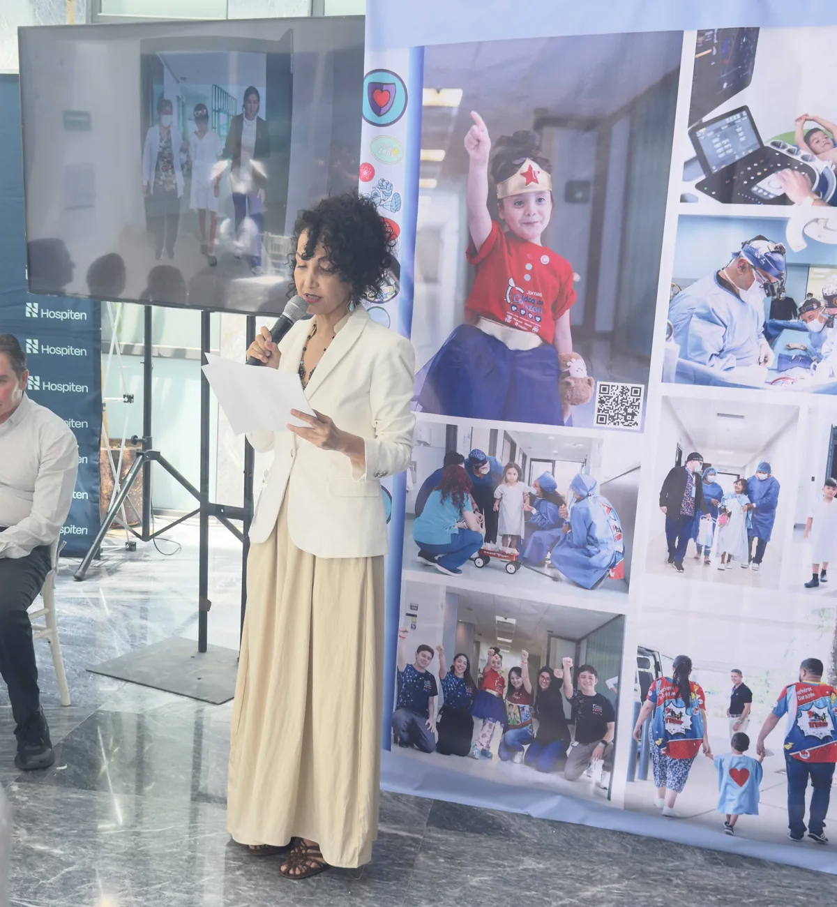 Woman in a white blazer reading from a paper while speaking into a microphone at an event, with a large collage banner behind her showing children and medical scenes