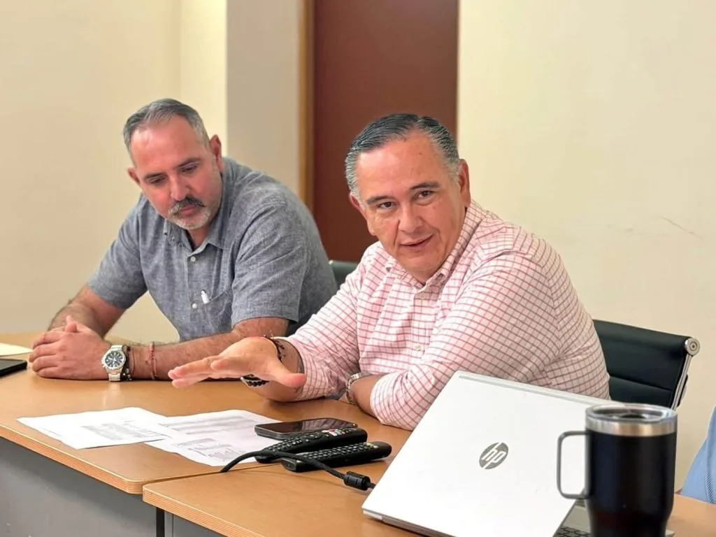 Two men sit at a conference table with papers, a silver HP laptop, remotes, and a black mug, one gesturing as they talk.