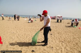 Volunteer on a sandy beach collects litter into a green bag during a cleanup event, with other volunteers in the background, sunny day.