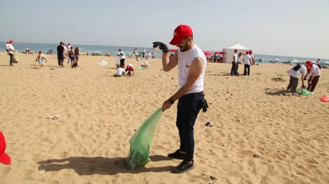 Volunteer on a sandy beach collects litter into a green bag during a cleanup event, with other volunteers in the background, sunny day.